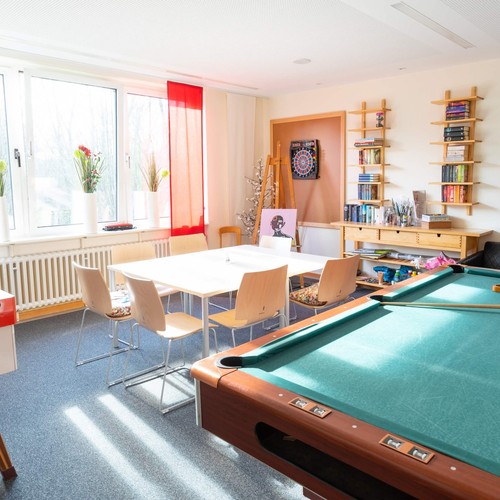 In einem hellen Zimmer mit großen Fenstern stehen ein Billiardtisch, eine Tischgruppe, Regale und eine Staffelei.