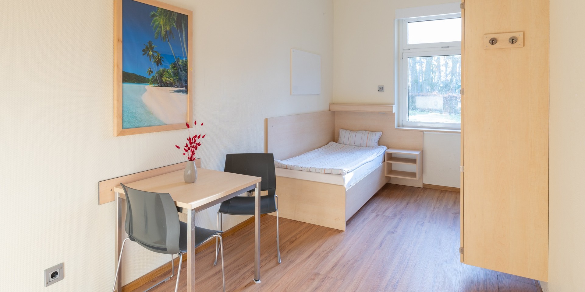 In einem hellen Patiententimmer stehen ein Bett mit weißer Bettwäsche, eine Tischgruppe und ein Schrank. An der Wand hängt ein Bild mit Palmen.