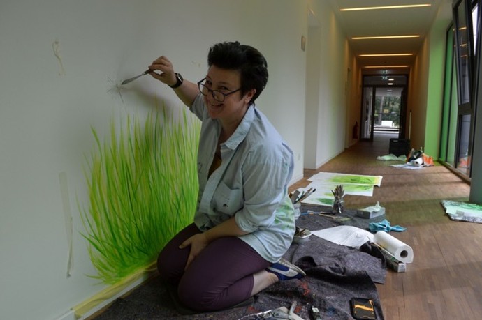 Das Bild zeigt eine Künstlerin, die zwischen Pinsel und Farbpaletten auf dem Boden sitzt und eine Blumenwiese an eine Wand malt.