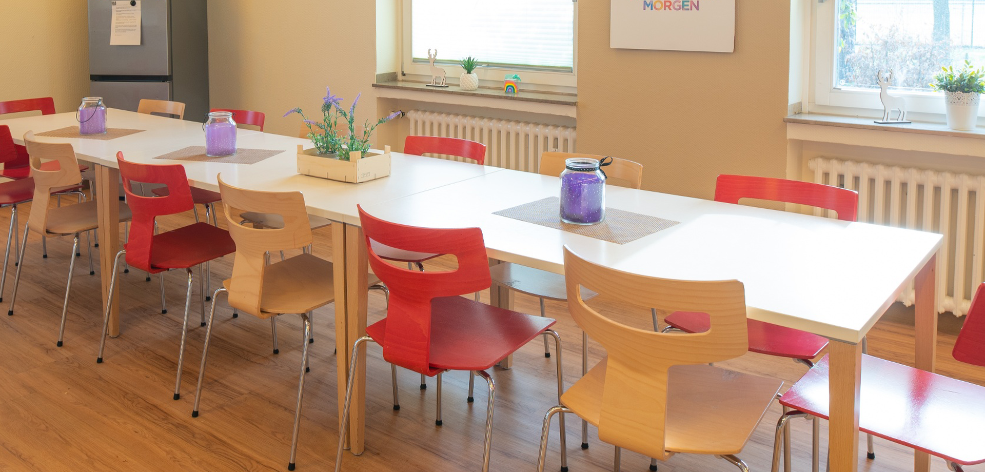 In einem Essraum steht ein großer Tisch mit Holzstühlen, die teilweise rot sind. Auf dem Tisch stehen violette Dekogläser und eine Pflanze.