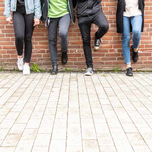 Das Bild zeigt vier Jugendliche, die nebeneinander, gelehnt an einer Backsteinmauer stehen. Nur ihre Unterkörper sind zu sehen.