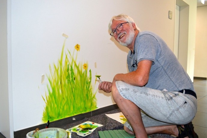 Das Bild zeigt einen Künstler, der eine Blumenwiese an eine Wand mit einem Pinsel malt.