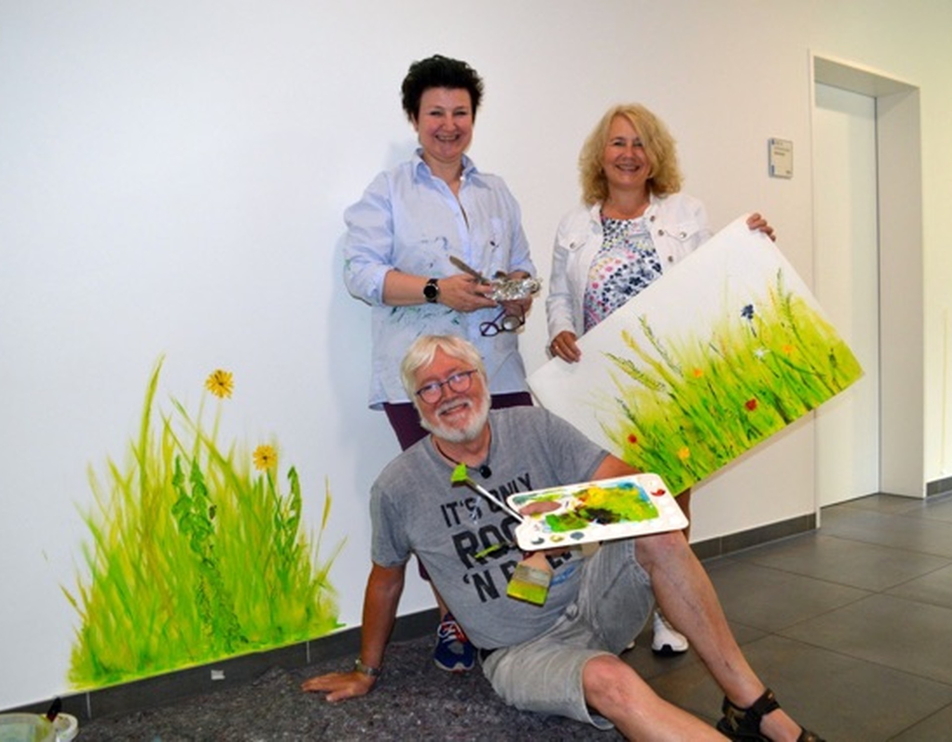 Das Bild zeigt drei Personen in der Klinik, darunter eine Künstlerin und ein Künstler. Die Personen halten ein Bild mit Blumen.