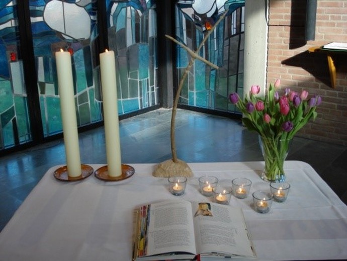 Auf einem Tisch, der in einer Kirche steht, stehen Kerzen, Tuplen und ein Kreuz. Auf der vorderen Hälfte des Tisches liegt ein aufgeschlagenes Buch.