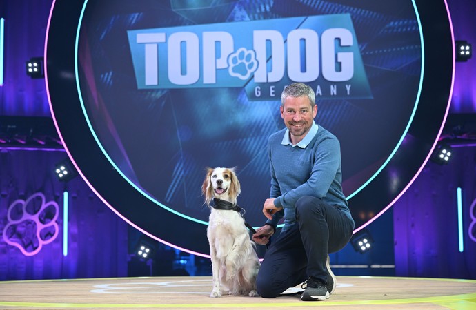 Das Bild zeigt einen Mann und einen Hund bei der Fernsehshow Top Dog. Die beiden stehen nebeneinander und schauen in die Kamera