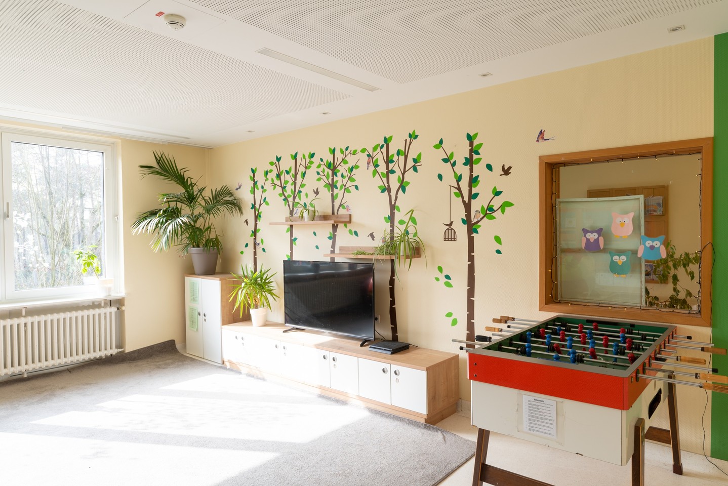 In dem Aufenthaltsraum stehen ein Kickertisch, ein Fernseher und eine Pflanze. Die Wände werden von Wandbildern geschmückt, die Bäume und Vögel zeigen