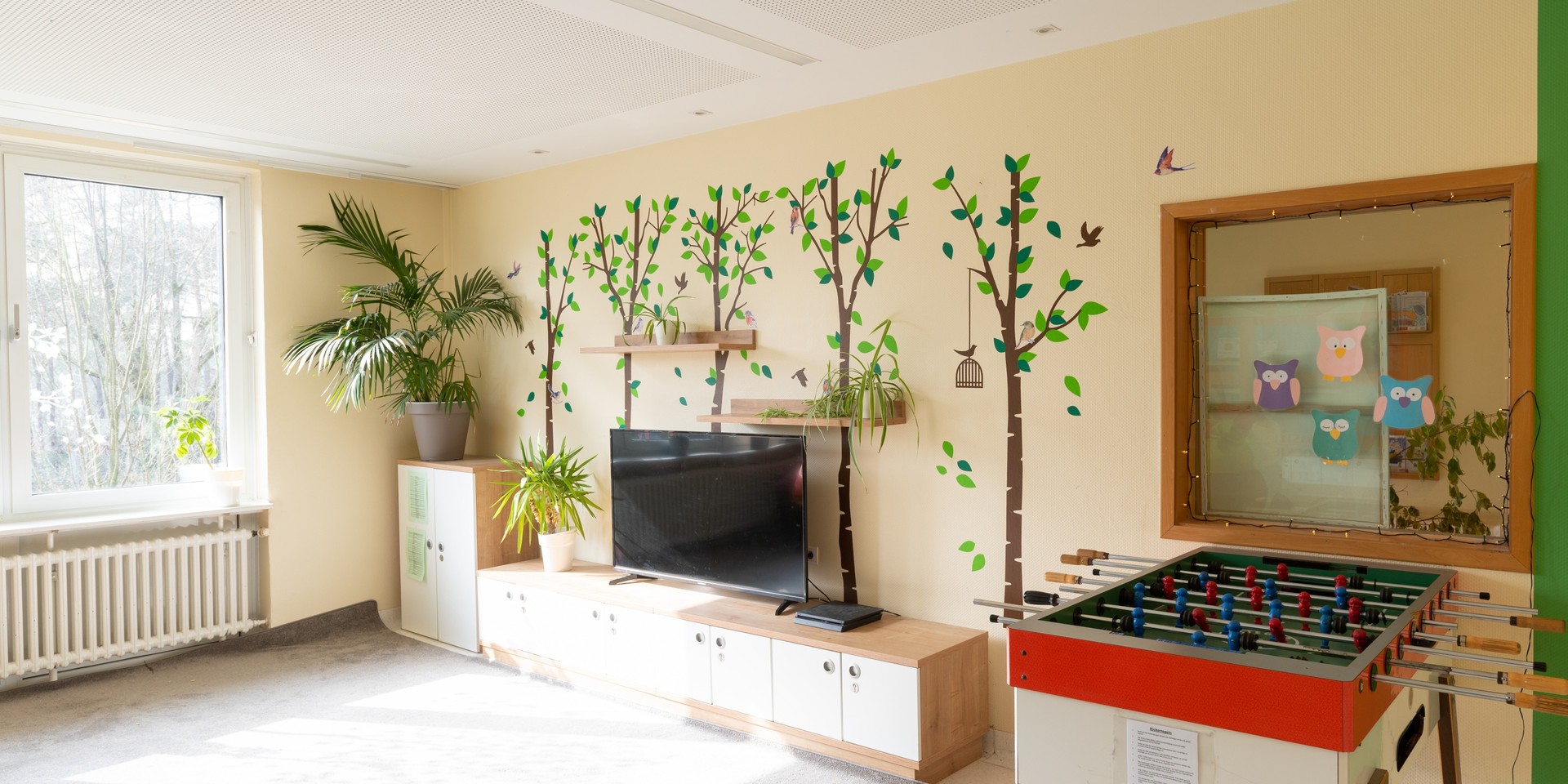 In dem Aufenthaltsraum stehen ein Kickertisch, ein Fernseher und eine Pflanze. Die Wände werden von Wandbildern geschmückt, die Bäume und Vögel zeigen