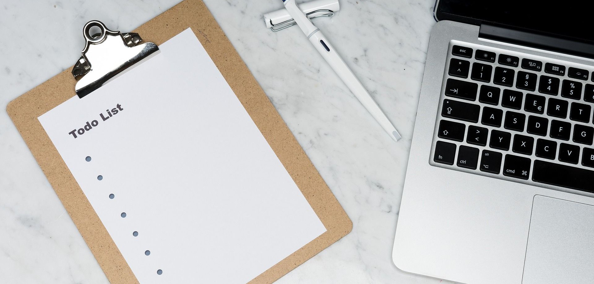 Das Bild zeigt ein Blatt auf einem Klemmbrett, auf dem "To Do List" steht, einen Füller und einen silbernen Laptop, die auf einem Marmortisch liegen.
