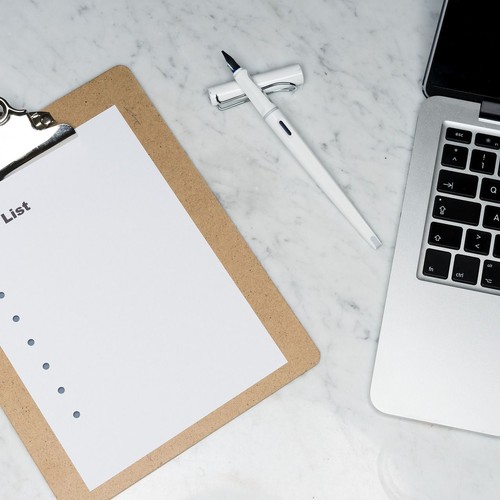 Das Bild zeigt ein Blatt auf einem Klemmbrett, auf dem "List" steht, einen weißen Füller und einen silbernen Laptop, die auf einem Marmortisch liegen.