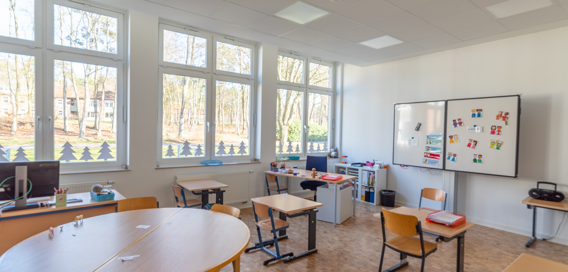 In einem hellen Klassenzimmer stehen Tische und Stühle, auf denen Stifte und Bastelmaterialien liegen. Durch die Fenster sieht man das grüne Gelände.