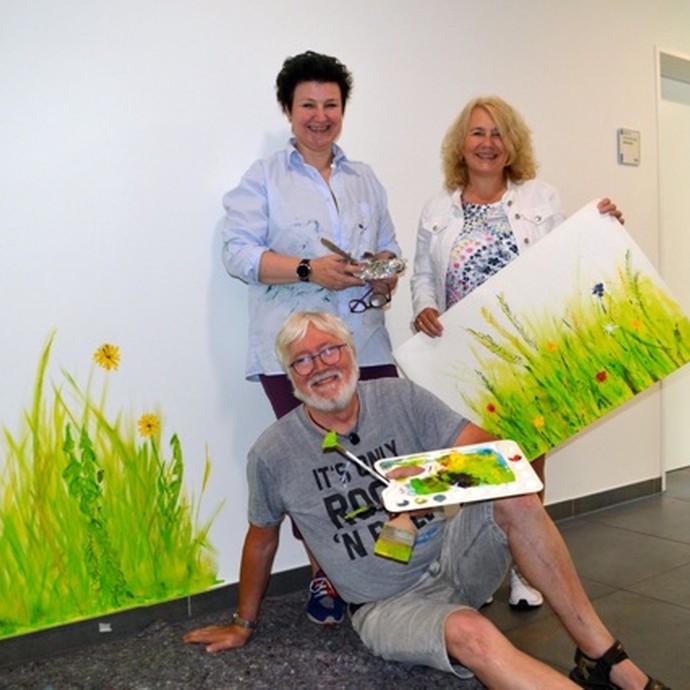 Das Bild zeigt eine Mitarbeiterin, einen Künstler und eine Künstlerin. Die Personen halten ein Bild mit einer Blumenwiese in die Kamera- (öffnet vergrößerte Bildansicht)