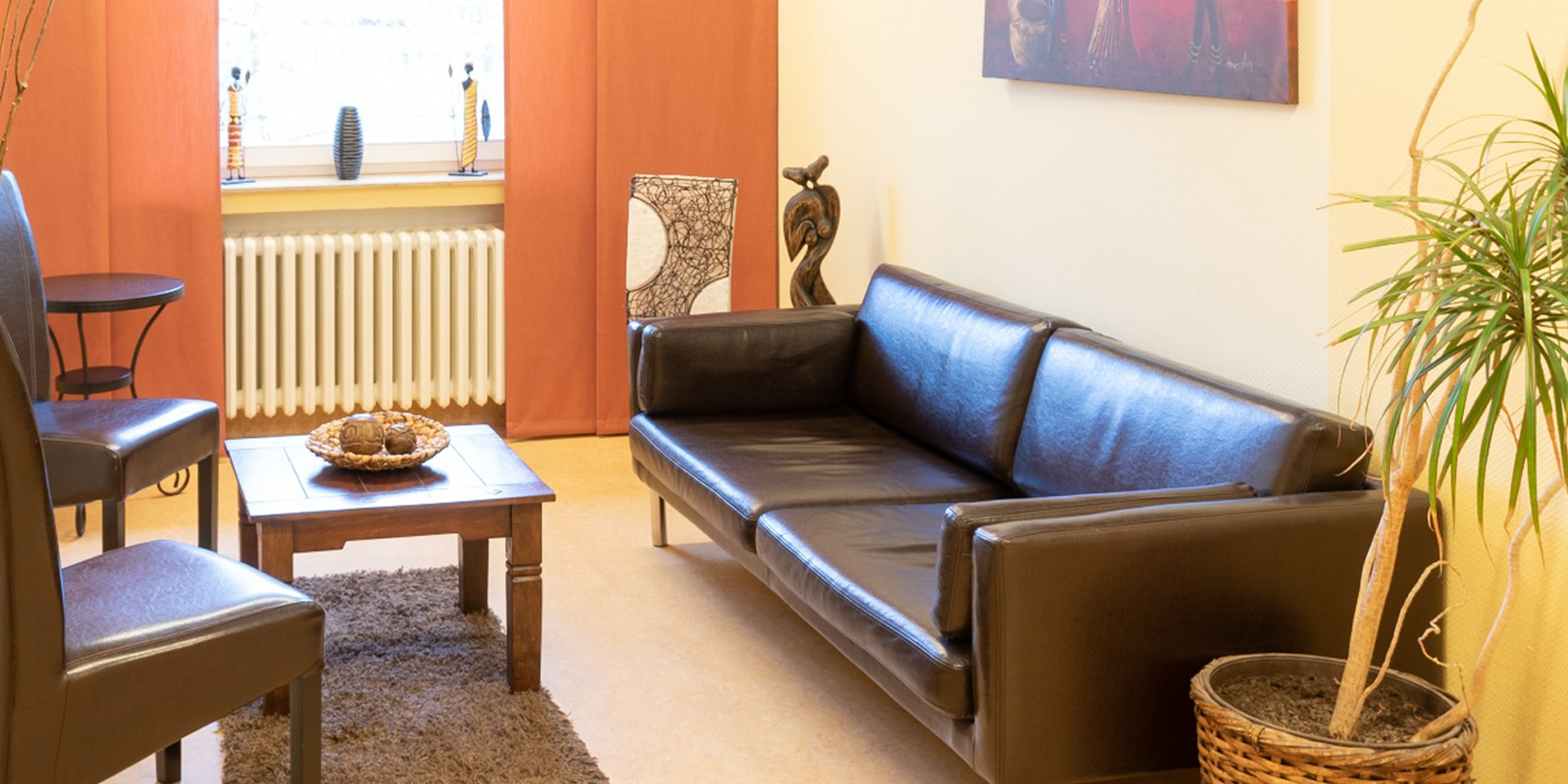 In dem warm gestalteten Therapieraum stehen ein braunes Ledersofa und braune Lederstühle sowie ein brauner Teppich und eine grüne Pflanze.