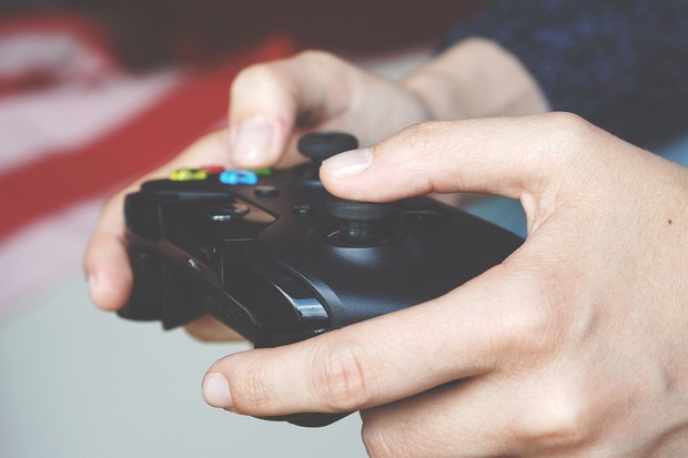 Das Bild zeigt eine Hand mit einem Playstation-Controller. Ein Jugendlicher spielt Playstation.