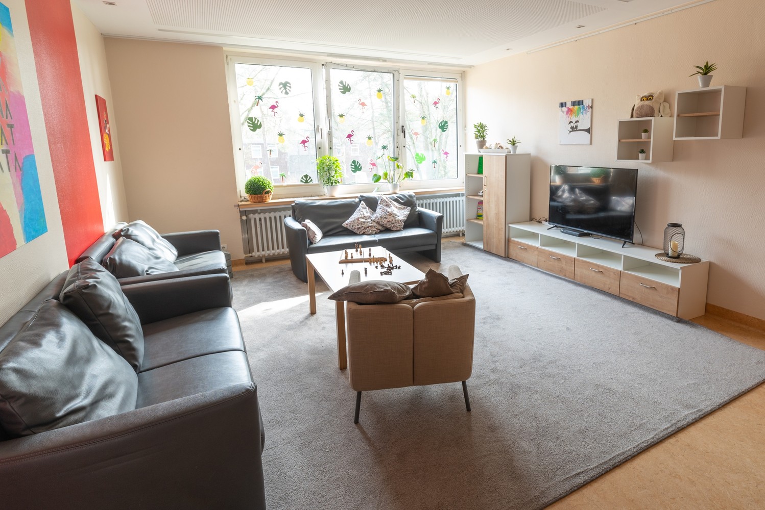 Das Bild zeigt einen hellen, großen Raum mit mehreren Sofas, einem grauen Teppich und einem Fernseher.