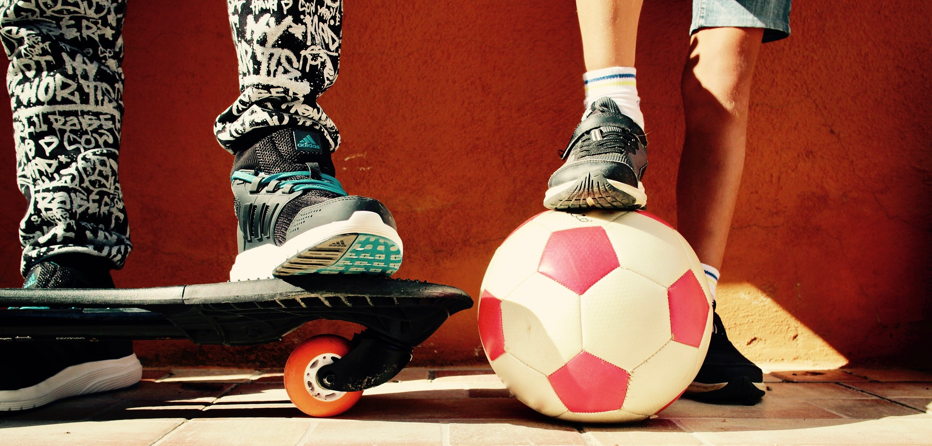 Zwei Jugendliche (man sieht nur ihre Beine) stehen nebeneinander, mit einem Ball und einem Skatebord.